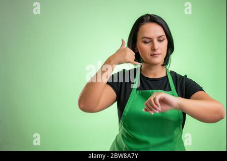 Cuoca con grembiule verde e t-shirt nera, mostrandomi un gesto di chiamata, guardando l'orologio isolato su sfondo verde Foto Stock