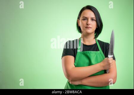 Cuoca con grembiule verde e t-shirt nera, che guarda sicuro con il coltello in mano isolato su sfondo verde Foto Stock