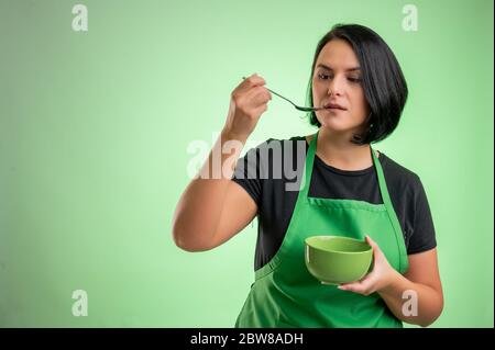 Cuoca con grembiule verde e t-shirt nera, assaggia la zuppa, tieni la ciotola in mano isolata su sfondo verde Foto Stock