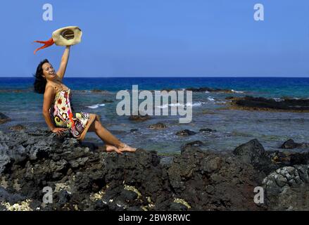 Bella donna hawaiana si siede su una spiaggia rocciosa della costa di Kohala e ondeggia un benvenuto. Indossa una spogliatrice e svita un cappello di paglia con colorfu Foto Stock