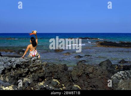 Bella donna hawaiana si siede su una spiaggia rocciosa della costa di Kohala e ondeggia un benvenuto. Indossa una spogliatrice e svita un cappello di paglia con colorfu Foto Stock