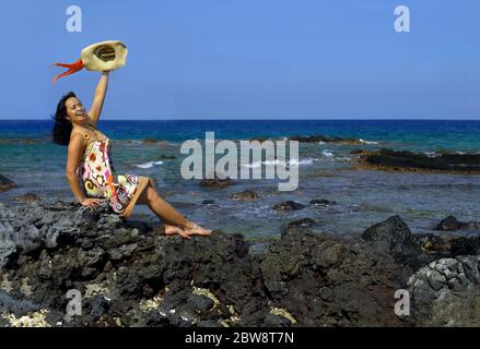 Bella donna hawaiana si siede su una spiaggia rocciosa della costa di Kohala e ondeggia un benvenuto. Indossa una spogliatrice e svita un cappello di paglia con colorfu Foto Stock