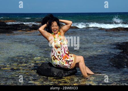 Donna hawaiana si siede su una roccia in acque poco profonde e celebra gettando i capelli e posando. Baia di Anaehomalu sulla costa di Kohala della Grande Isla Foto Stock