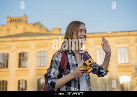 Giovane fotografo hipster che scatta foto, cammina in città e ride. Ritratto di felice ragazza turistica con zaino e fotocamera gialla Foto Stock