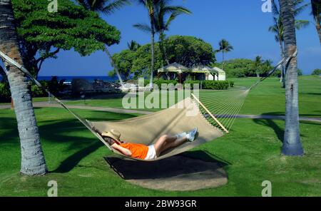 La donna anziana si trova in un'amaca tra due palme in un resort sulla costa di Kohala della Big Island delle Hawaii. Il suo cappello di paglia si stende su di lei Foto Stock