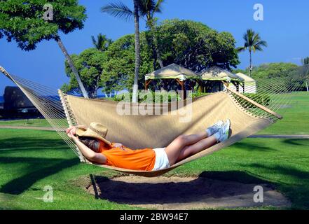 La donna anziana si trova in un'amaca tra due palme in un resort sulla costa di Kohala della Big Island delle Hawaii. Il suo cappello di paglia si stende su di lei Foto Stock