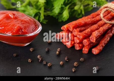 Primo piano di kabanos o cabanossi di salsiccia affumicata secca e ketchup di pomodoro su un asse da servizio in pietra nera. Tradizionale delicatezza di carne lucidata. Foto Stock