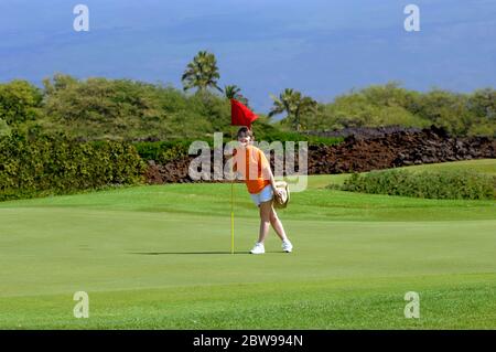 Donna si erge oltre al buco segnato da una bandiera rossa su un campo da golf sulla Big Island delle Hawaii. Mauna Loa sorge sullo sfondo dietro il campo da golf. Foto Stock