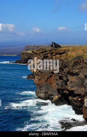 Lone camper pesca South Point, sulla Big Island delle Hawaii. Le onde si tuffano contro le rocce alla base di una ripida scogliera. Foto Stock