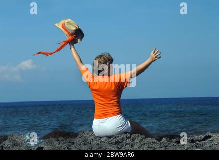 La donna anziana si siede su rocce laviche nere sulla costa frastagliata della costa di Kona della Big Island delle Hawaii. Sta sventolando il suo cappello di paglia e la sc arancione Foto Stock