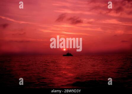 La barca naviga attraverso il riflesso di un sole rosso al tramonto. Solitaria barca su un vasto oceano di colore. Foto Stock