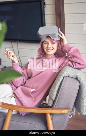 Felice hipster teen girl indossare vr visore tenere controller guardare la telecamera in sedia. Foto Stock