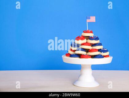 Piedistallo in porcellana bianca con vivaci e colorati biscotti allo zucchero ghiacciati di dimensioni patriotiche con piccola bandiera americana sulla parte superiore seduta su un tavolo di legno chiaro. Foto Stock