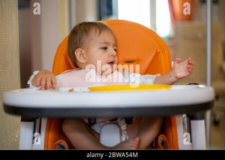 Cute bambina caucasica sedersi in poltrona arancione dopo aver mangiato con briciole sul viso e guarda a portata di mano su briciole da tavolo e una piastra di plastica arancione Foto Stock