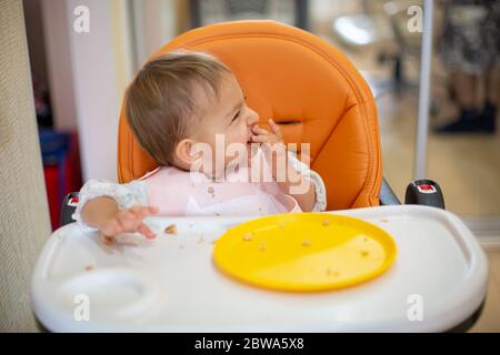 il bambino carino si siede su una seggioletta arancione con un tavolo con le mani e il viso sporchi, guarda via e ride. sul tavolo ci sono briciole e un piatto arancione. chiudi Foto Stock