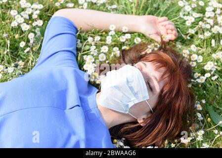 Ragazza sul prato fiorito di margherite con maschera chirurgica. Una ragazza in un giardino fiorito è sdraiata sull'erba Foto Stock