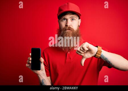 Redhead irlandese delivery man con barba che tiene in mano smartphone che mostra app sullo schermo con viso arrabbiato, segno negativo che mostra dispiacere con pollici giù, rifiutare Foto Stock