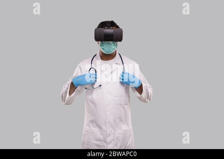 Medico che indossa occhiali VR, maschera medica e guanti che tiene stetoscopio isolato. Medicina in linea del medico dell'uomo indiano Foto Stock