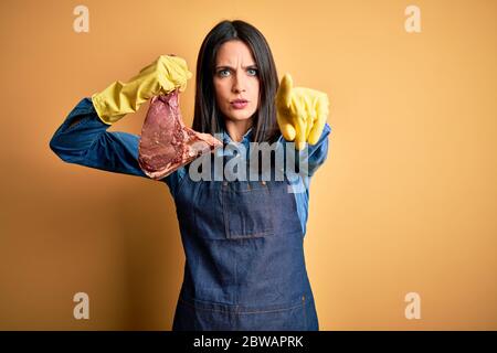 Giovane macellaio donna che indossa grembiule tenendo la bistecca di manzo crudo su sfondo giallo isolato puntando con il dito alla macchina fotografica e a voi, segno di mano, pos Foto Stock