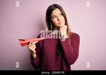 Giovane bella ragazza che tiene l'aereo di carta in piedi su sfondo rosa isolato faccia seria che pensa alla domanda, idea molto confusa Foto Stock