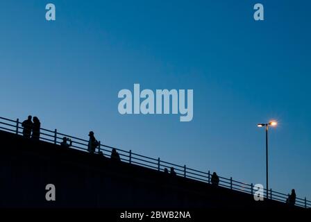 Silhouette di persone che camminano attraverso il ponte di Waterloo, fotografate al tramonto con un lampione illuminato. South Bank, Londra, Regno Unito. Orizzontale. Foto Stock