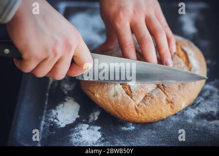 Un uomo taglia con un coltello affilato pane di grano fatto in casa appena fatto sdraiato su una teglia scura cottura a casa. Foto Stock