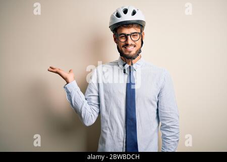 Giovane uomo d'affari che indossa occhiali e casco da bici in piedi su isolato panettaio bianco sorridente allegro presentazione e puntando con palmo di mano lo Foto Stock