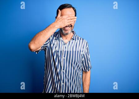 Uomo di mezza età che indossa camicia a righe e occhiali da sole su sfondo blu isolato sorridendo e ridendo con mano sulla faccia che copre gli occhi per surpr Foto Stock