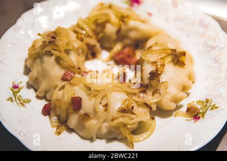 Tradizionali gnocchi polacchi fatti in casa - pierogis - su piatto di porcellana servito con cipolla fritta e pancetta. Primo piano di gnocchi di patate Foto Stock