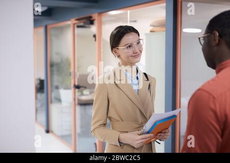 Vita su ritratto di giovane donna d'affari contemporanea che parla con il collega afroamericano e sorridente allegro, mentre si trova in ufficio sala, poliziotto Foto Stock