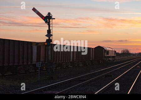 DB Cargo Rail locomotiva UK classe 66 66161 trasporto di un treno merci che trasporta minerale di ferro che passa i segnali semaforici a Barnetby, Lincs, al tramonto Foto Stock