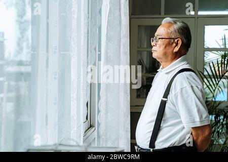 Primo piano di un vecchio uomo penoso con occhiali che guardano fuori dalla finestra sentirsi soli. Anni 70 anziani asiatici pensionati scomparsi figli e moglie. Foto Stock