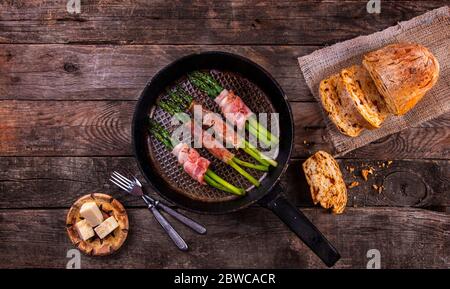 Asparagi verdi arrostiti avvolti in pancetta in padella vecchia, formaggio e pane fatto in casa con pomodori secchi su sfondo rustico di legno. Vista dall'alto. Copia sp Foto Stock