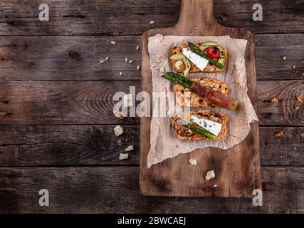 Pane tostato fatto in casa servito con asparagi verdi avvolti in pancetta, formaggio, fette di avocado, ciliegia di pomodoro alla griglia e noci su legno rustico antico Foto Stock