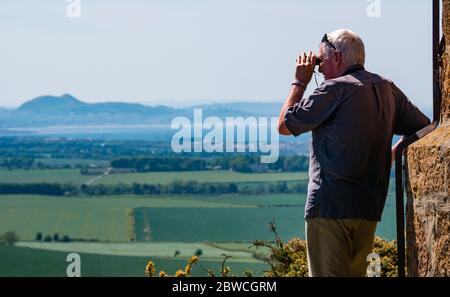 East Lothian, Scozia, Regno Unito, 31 maggio 2020. Regno Unito Meteo: Più caldo tempo in tutta la contea, ma con il sole frizzante. Un uomo anziano guarda attraverso il binocolo dalla cima di Byres Hill con il profilo distintivo di Arthur's Seat in lontananza Foto Stock