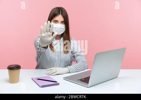 Divieti di quarantena. Donna lavoro in ufficio seduto sano sicuro con maschera igienica viso e guanti protettivi, mostrando gesto di stop. Lavorare in auto-isolati Foto Stock