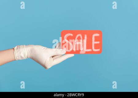 Profilo Vista laterale primo piano della mano umana in guanti chirurgici bianchi che tengono e mostrano etichetta vendita rossa. Interno, studio shot, isolato su sfondo blu. Foto Stock