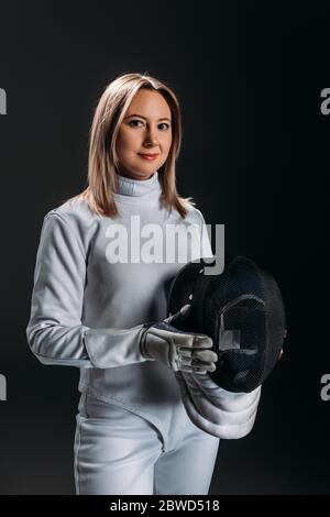 Attraente finitore in tuta scherma che tiene la maschera e guardando la fotocamera isolata su nero Foto Stock