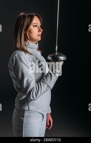 Vista laterale della donna in tuta recinzione che tiene la pinza isolata su nero Foto Stock