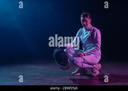 Bella finitrice in tuta scherma che tiene maschera e pinza su sfondo nero con illuminazione Foto Stock