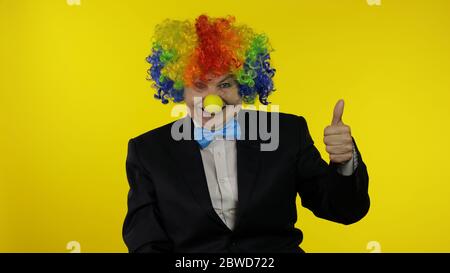 Il vecchio clown caucasico femminile senior in parrucca colorata e cravatta blu mostra pollice su. Donna anziana che si diverte, sorridente. Spazio di copia. Halloween. Sfondo giallo Foto Stock