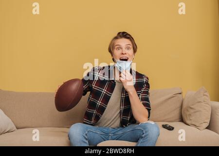 eccitato giovane uomo in maschera medica che tiene il calcio americano mentre guarda il campionato in soggiorno Foto Stock