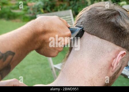 Un uomo tenta di tagliare i capelli di un altro uomo durante il blocco del coronavirus quando i parrucchieri sono chiusi, lasciando la coda di un ratto giù un lato della testa Foto Stock