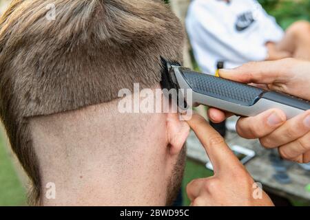 Un uomo tenta di tagliare i capelli di un altro uomo durante il blocco del coronavirus quando i parrucchieri sono chiusi, lasciando la coda di un ratto giù un lato della testa Foto Stock