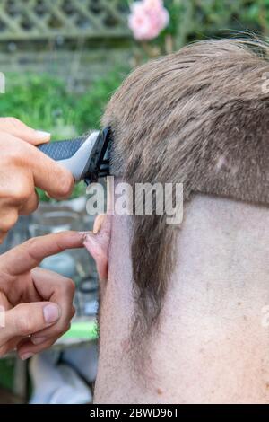 Un uomo tenta di tagliare i capelli di un altro uomo durante il blocco del coronavirus quando i parrucchieri sono chiusi, lasciando la coda di un ratto giù un lato della testa Foto Stock