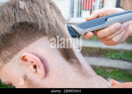 Un uomo tenta di tagliare i capelli di un altro uomo durante il blocco del coronavirus quando i parrucchieri sono chiusi, lasciando la coda di un ratto giù un lato della testa Foto Stock