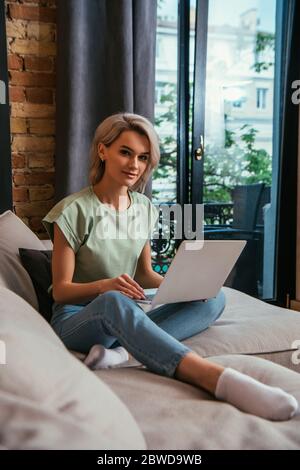 giovane donna che sorride alla fotocamera mentre si siede sul divano vicino alla finestra e si usa il computer portatile Foto Stock