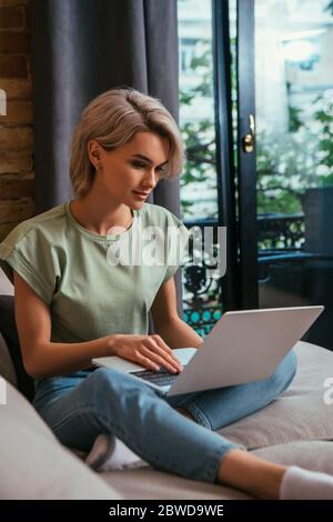 donna giovane concentrata seduta sul divano vicino alla finestra e utilizzando il computer portatile Foto Stock