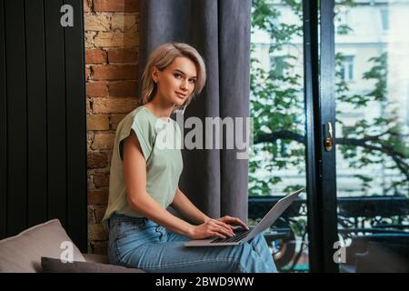 felice giovane donna che guarda la fotocamera mentre usa il laptop vicino alla finestra di casa Foto Stock