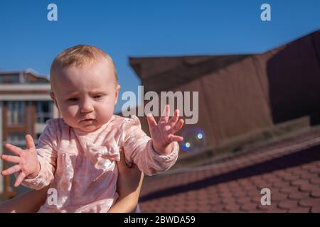 Primo piano di cute baby girl indossare adorabile vestito sul tetto, bolle di sapone galleggianti intorno, bambina cercando di prendere le bolle Foto Stock